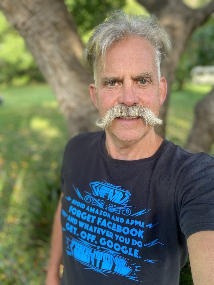 Roly sporting a large moustache wearing a black T-Shirt with the words WFMU,  avoid amazon and apple forget Facebook and whatever you do get off Google, and ending with the words TECHTONIC.